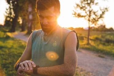 Young sportsman examining time on smart watch standing in park - SBAF00054