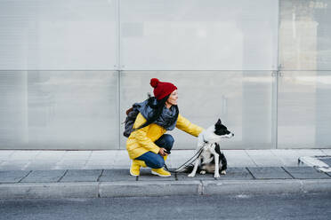 Woman looking away while crouching with dog on footpath - EBBF01198