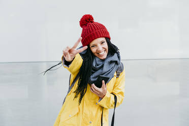 Woman smiling while doing peace sign holding smart phone while standing against wall - EBBF01203