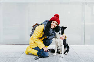 Lächelnde Frau, die ihren Hund umarmt, während sie auf dem Gehweg vor einer Mauer kniet - EBBF01205