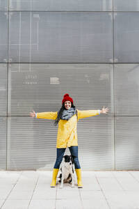 Glückliche Frau mit ausgestreckten Armen, die mit ihrem Hund auf einem Fußweg vor einem Gebäude steht - EBBF01210