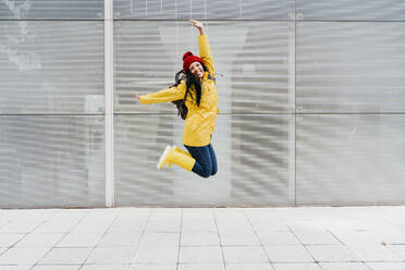 Cheerful woman jumping with arms raised on footpath against building exterior - EBBF01211