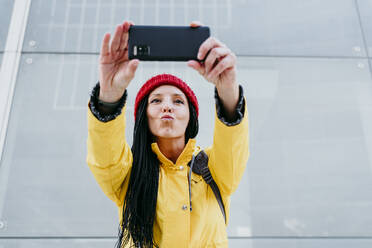 Frau, die ein Selfie mit ihrem Smartphone macht, während sie an einer grauen Wand steht - EBBF01217