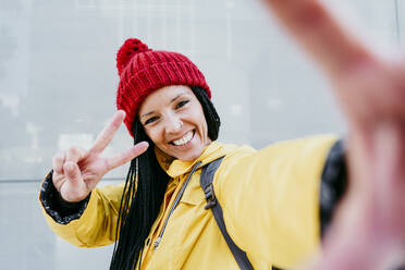 Smiling woman doing peace sign while standing against gray wall during autumn - EBBF01218