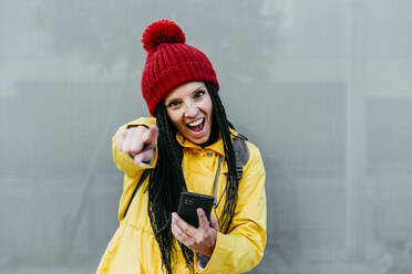 Woman pointing with mouth open while holding smart phone standing against gray wall - EBBF01224