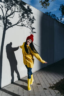 Lächelnde Frau im Regenmantel steht auf dem Fußweg an der Mauer im Schatten eines Baumes - EBBF01255