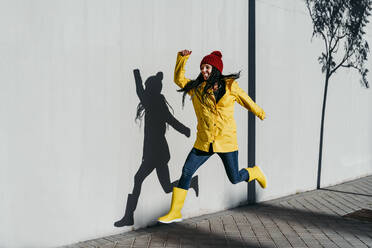 Cheerful woman wearing raincoat jumping on footpath by wall - EBBF01256