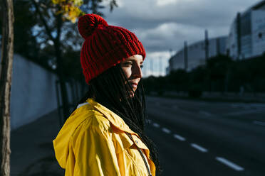 Nachdenkliche Frau mit Strickmütze steht bei Sonnenuntergang auf der Straße - EBBF01257