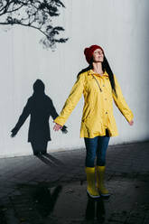 Woman wearing raincoat standing on rain puddle against shadow wall - EBBF01261