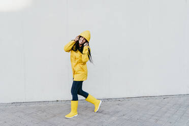 Smiling woman wearing raincoat standing on footpath against gray wall - EBBF01266