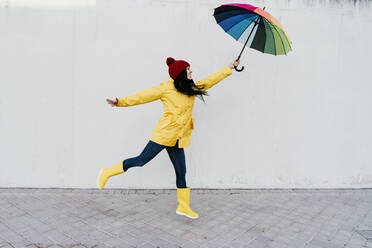 Frau mit ausgestreckten Armen, die einen bunten Regenschirm hält, während sie auf einem Fußweg an der Wand steht - EBBF01269