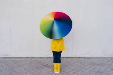 Woman holding colorful umbrella standing in front of gray wall on footpath - EBBF01271