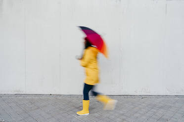 Woman walking with colorful umbrella on footpath by wall - EBBF01273