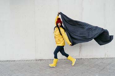 Woman wearing raincoat holding blanket while walking by wall on footpath - EBBF01278