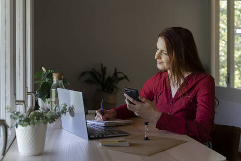 Female doctor with mobile phone writing in book while listening to video call on laptop at home - AFVF07449