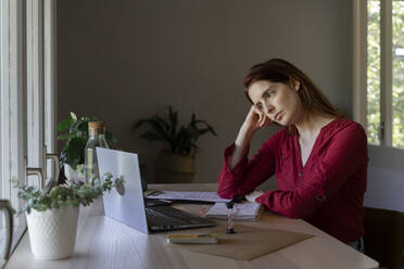 Doctor with head in hands listening to video call on laptop while sitting at home - AFVF07452