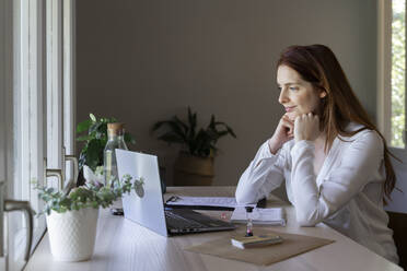 Weiblicher Arzt mit Hand am Kinn, der zu Hause sitzend einen Videoanruf auf dem Laptop hört - AFVF07456