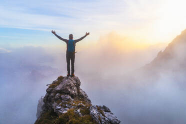 Pensive male hiker with arms outstretched standing on mountain peak during sunrise at Bergamasque Alps, Italy - MCVF00647