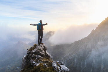 Pensive hiker with arms outstretched standing on mountain peak during sunrise at Bergamasque Alps, Italy - MCVF00648