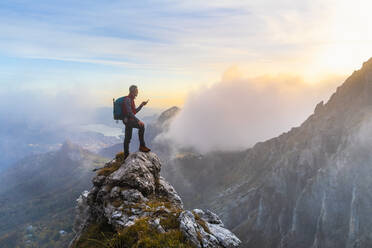 Pensive hiker using smart phone on mountain peak during sunrise at Bergamasque Alps, Italy - MCVF00649
