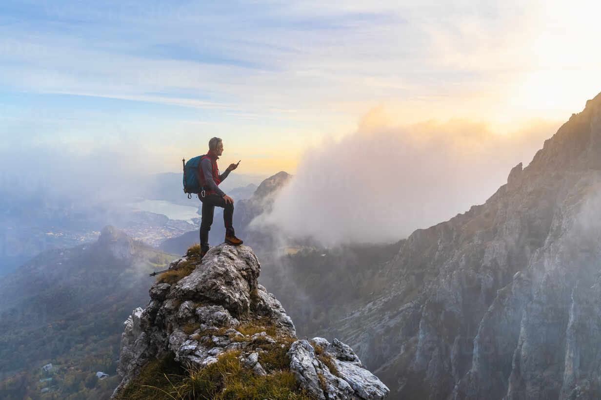 Pensive hiker using smart phone on mountain peak during sunrise at