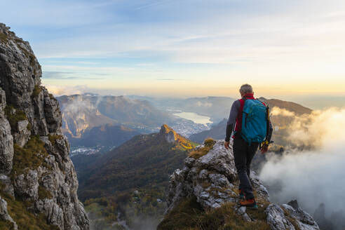 Nachdenklicher männlicher Wanderer bei Sonnenaufgang auf einem Berggipfel in den Bergamasker Alpen, Italien - MCVF00650