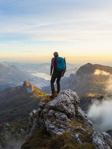 Nachdenklicher Wanderer mit Rucksack auf einem Berggipfel bei Sonnenaufgang in den Bergamasker Alpen, Italien - MCVF00651