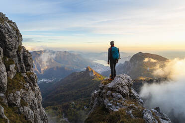 Nachdenklicher Wanderer, der die Aussicht betrachtet, während er bei Sonnenaufgang auf einem Berggipfel in den Bergamasker Alpen, Italien, steht - MCVF00652