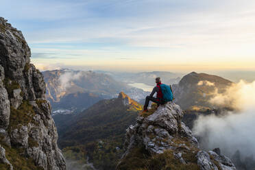 Nachdenklicher Wanderer, der die Aussicht betrachtet, während er bei Sonnenaufgang auf einem Berggipfel in den Bergamasker Alpen, Italien, sitzt - MCVF00653