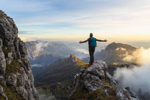 Nachdenklicher Wanderer mit ausgestreckten Armen auf einem Berg stehend während des Sonnenaufgangs in den Bergamasker Alpen, Italien - MCVF00654