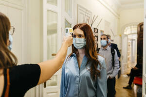Female with infrared thermometer checking businesswoman's temperature in corridor - JRFF04797
