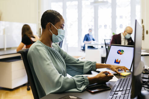 African businesswoman with safety mask working at office desk - JRFF04808