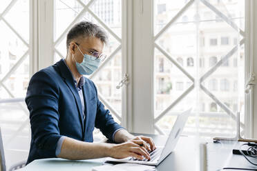 Male entrepreneur with protective mask working on laptop against window in office - JRFF04811