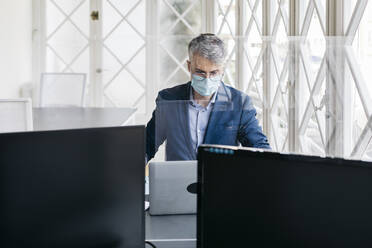 Männlicher Unternehmer mit Schutzmaske bei der Arbeit an einem Laptop in einem Büro, gesehen durch eine Glasscheibe - JRFF04812