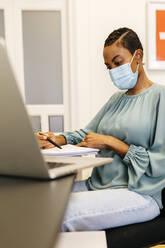 African businesswoman with safety mask reading papers while sitting in office - JRFF04814