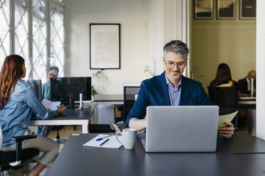 Lächelnder Geschäftsmann arbeitet am Laptop am Schreibtisch im Büro - JRFF04841