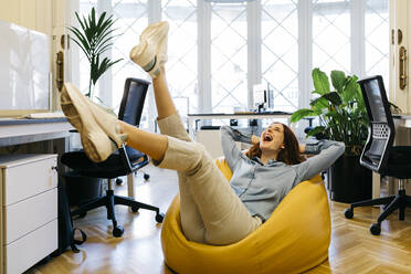 Happy businesswoman with hands behind head relaxing on bean bag at workplace - JRFF04866