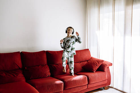 Happy boy listening to music through headphones while dancing on sofa at home - XLGF00730