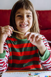 Happy girl with paintbrush showing toothed gap at home - XLGF00735