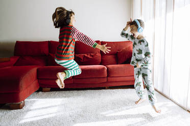 Männliche und weibliche Geschwister tragen Kopfhörer, während sie gegen das Sofa im Wohnzimmer tanzen - XLGF00744