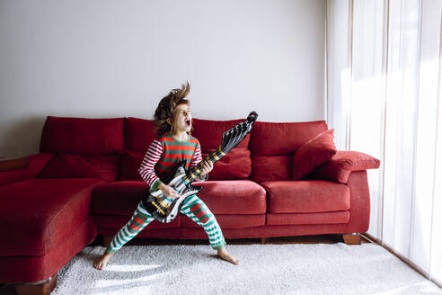 Happy girl playing guitar against sofa in living room - XLGF00747