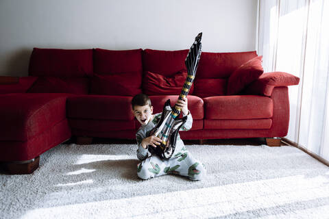 Glücklicher Junge spielt Gitarre, während er gegen das Sofa im Wohnzimmer kniet, lizenzfreies Stockfoto