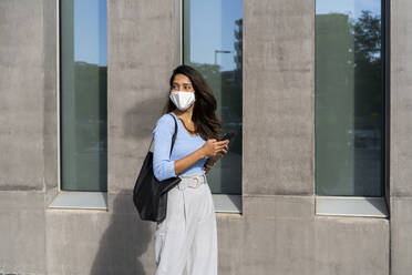 Junge Frau mit Gesichtsschutz, die während COVID-19 an einem Gebäude vorbeigeht - AFVF07460