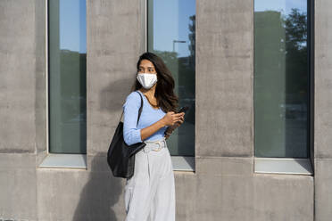 Young woman in protective face mask walking by building during COVID-19 - AFVF07460