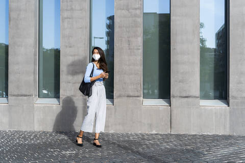 Geschäftsfrau mit Gesichtsmaske steht während der Coronavirus-Krise auf dem Gehweg vor einem Gebäude, lizenzfreies Stockfoto
