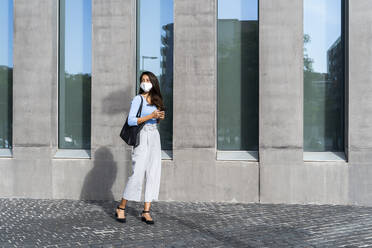 Businesswoman in face mask standing on footpath by building during coronavirus crisis - AFVF07461