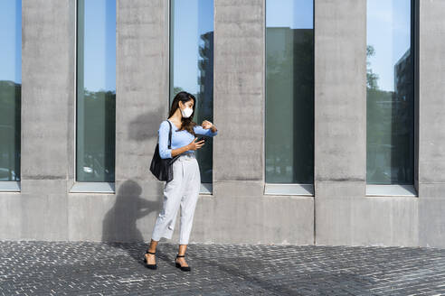 Businesswoman in face mask checking time on wristwatch while standing on footpath during pandemic - AFVF07462