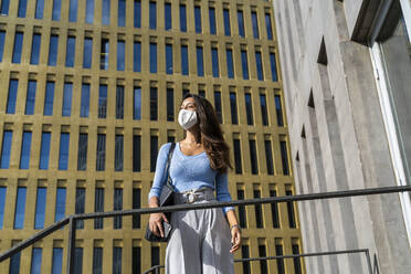 Junge Geschäftsfrau mit Gesichtsmaske schaut weg, während sie während des Ausbruchs des Coronavirus vor einem Bürogebäude steht - AFVF07464