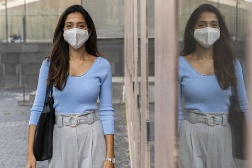 Young businesswoman in protective mask standing by glass window during pandemic - AFVF07467