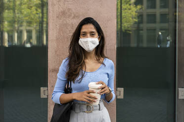 Junge Geschäftsfrau mit Schutzmaske, die einen Einweg-Kaffeebecher hält, während sie an einem Gebäude steht - AFVF07469